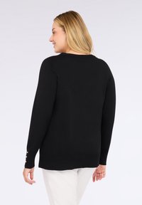 Žena s blond vlasy má na sobě černý svetr s dlouhým rukávem a zlatými knoflíky na manžetách, bílé kalhoty, stojí zády k nám a mírně se usmívá směrem doleva.