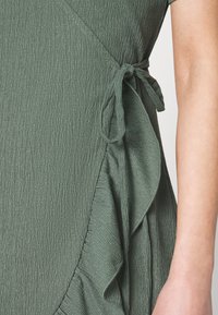 Vihreä teksturoitu mekko, jossa on röyhelöhelma ja solmimisnauha vyötäröllä, esittelee pehmeän, hieman ryppyisen kankaan.