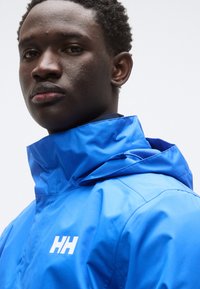 Μπλε αδιάβροχο μπουφάν με ψηλό γιακά, που διαθέτει λευκό λογότυπο και υφασμάτινη υφή, προσφέροντας ομαλή επιφάνεια και πρακτικό σχεδιασμό.