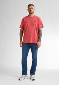 Koraal kleurig t-shirt met korte mouwen en een logo op de borst, gecombineerd met blauwe spijkerbroek en witte sportschoenen. Eenvoudig ontwerp, zachte textuur en een relaxte pasvorm.