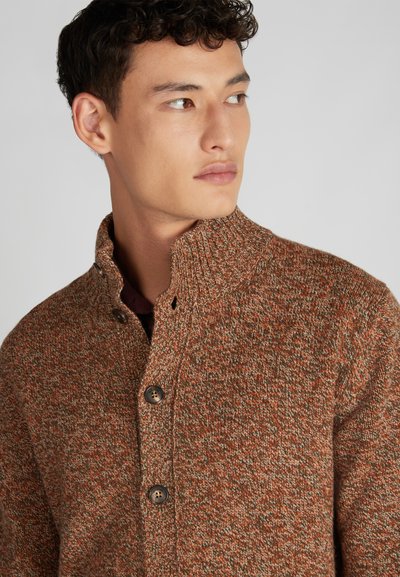 Sweter o teksturowanym splocie w ciepłych odcieniach pomarańczy i brązu, z wysokim kołnierzem i brązowymi guzikami. Zbliżenie na ko łnierz i detale swetra.