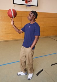 Mørkeblå t-shirt med et hvidt logo, beige bukser og hvide sneakers med grå detaljer, der balancerer en rød basketball i et gym-miljø.
