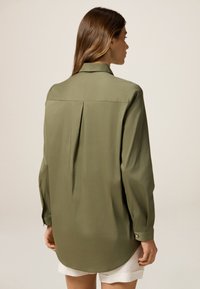 Camicia verde oliva a maniche lunghe con una piega sul retro, colletto sartoriale e polsini con bottoni. Tessuto liscio e orlo leggermente curvato.