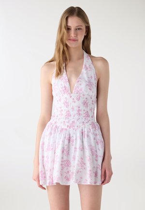 Młoda kobieta w białej sukience na szyję z różowymi kwiatowymi wzorami, stojąca na jednolitym jasnym tle.