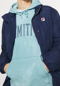 Marinblå pufferjacka över en ljusblå hoodie. Jackan har dragkedja fram och en logotypapplikation; hoodien har en tryckt textdesign.