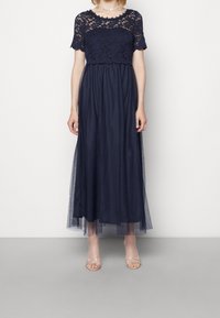 Marinblå maxiklänning med ett spetsöverdel och korta ärmar, en figurnära bodice och en svävande tylskirt. Accenter inkluderar genomskinlig överdel.