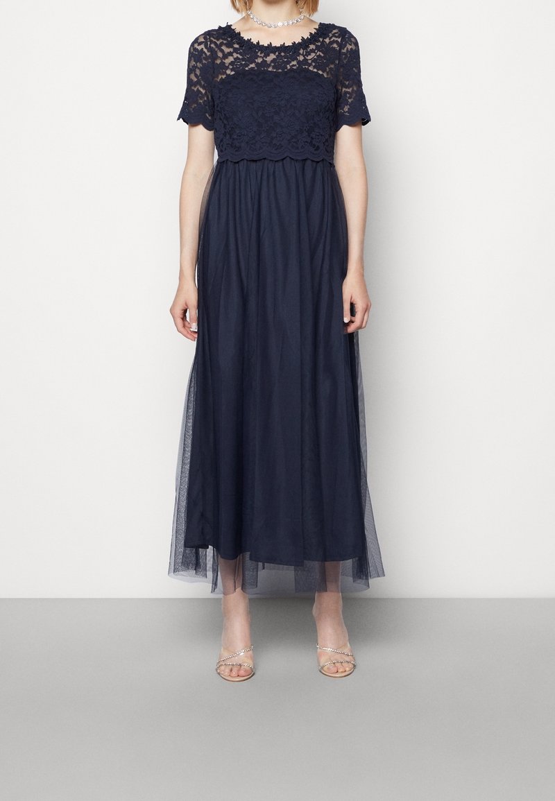 Marinblå maxiklänning med ett spetsöverdel och korta ärmar, en figurnära bodice och en svävande tylskirt. Accenter inkluderar genomskinlig överdel.