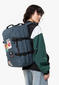 Young person wearing a green and black jacket and light jeans, carrying a large blue backpack with multiple colorful patches.