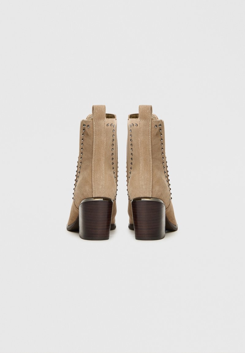 Beżowe zamszowe botki sięgające kostki z okrągłym noskiem, dekoracyjnymi ćwiekami po bokach i masywnym drewnianym obcasem.