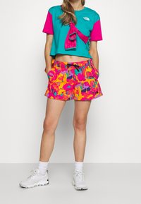 Donna che indossa una maglietta cropped color turchese e rosa, pantaloni shorts con stampa astratta colorata, calze bianche e sneakers bianche, in piedi contro uno sfondo bianco.