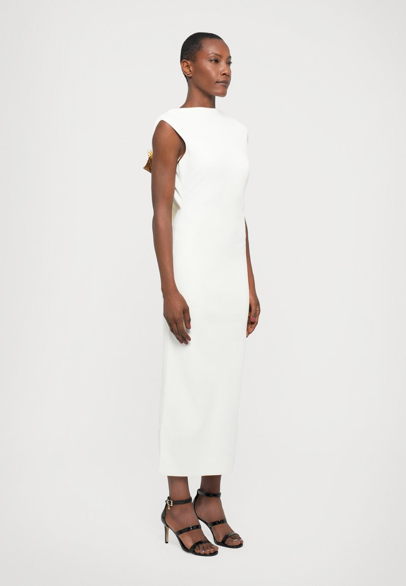 Robe blanche sans manches avec un col haut et une fente sur le côté, ornée de détails dorés. Portée avec des talons noirs à lanières.
