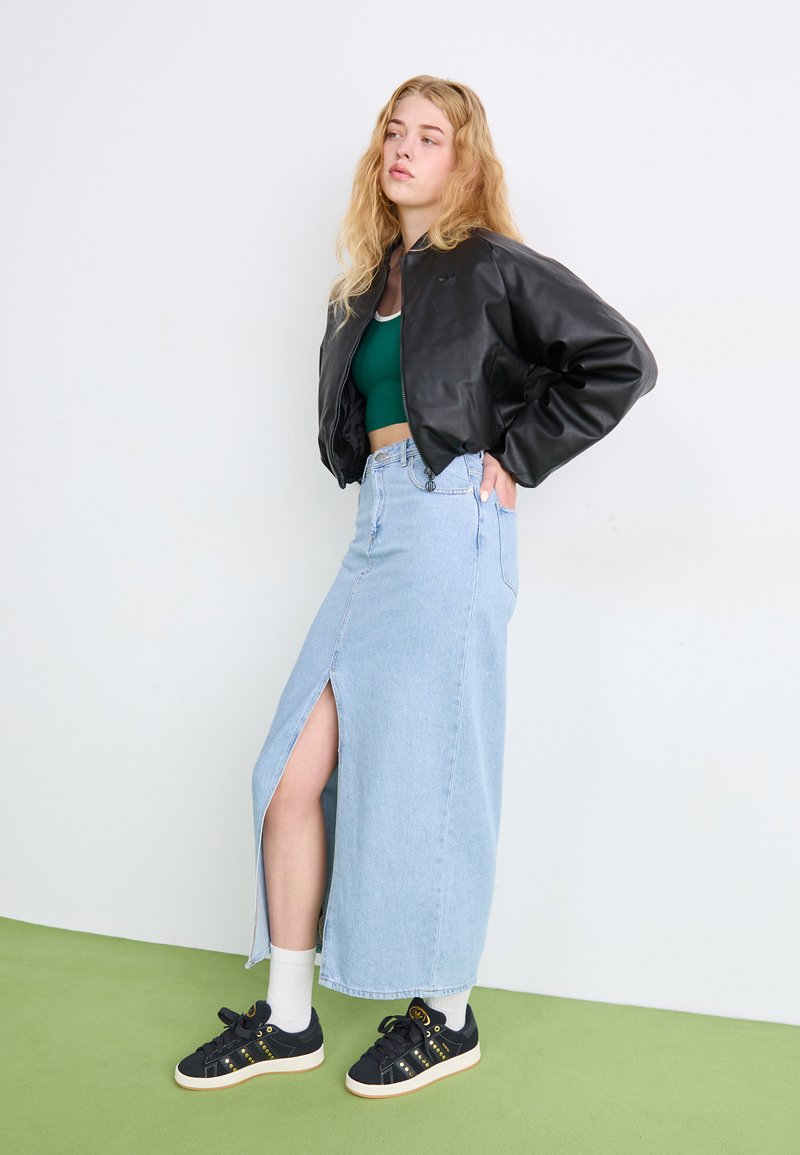 Mustmust nahast bomber-jakk, roheline lühike topp, helesinine denim maksiseelik esiseeliku lõhega ja mustad tenningud kuldsete aktsentidega rohelisel põrandal.