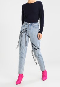 Jeans a vita alta blu chiaro con testo ricamato nero e borchie lunghe, abbinati a un maglione blu navy e stivali con tacco rosa.