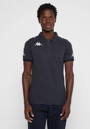 Uomo in piedi di fronte a noi, indossa una polo scura a maniche corte con loghi bianchi sulle spalle e sulle maniche e pantaloni scuri.