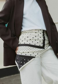 Patterned scarf featuring black dots on a cream background, with triangular shapes and a black border; paired with a black blazer and white pants.