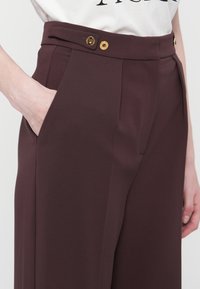 Personne portant un pantalon taille haute marron foncé avec deux boutons dorés à la taille et une main dans la poche gauche.