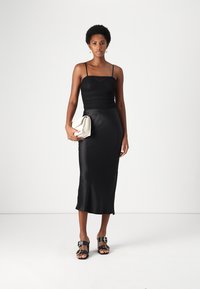 Robe midi en satin noir avec des bretelles spaghetti, un corsage ajusté et une jupe fluide. Le modèle tient une pochette crème et porte des sandales noires.