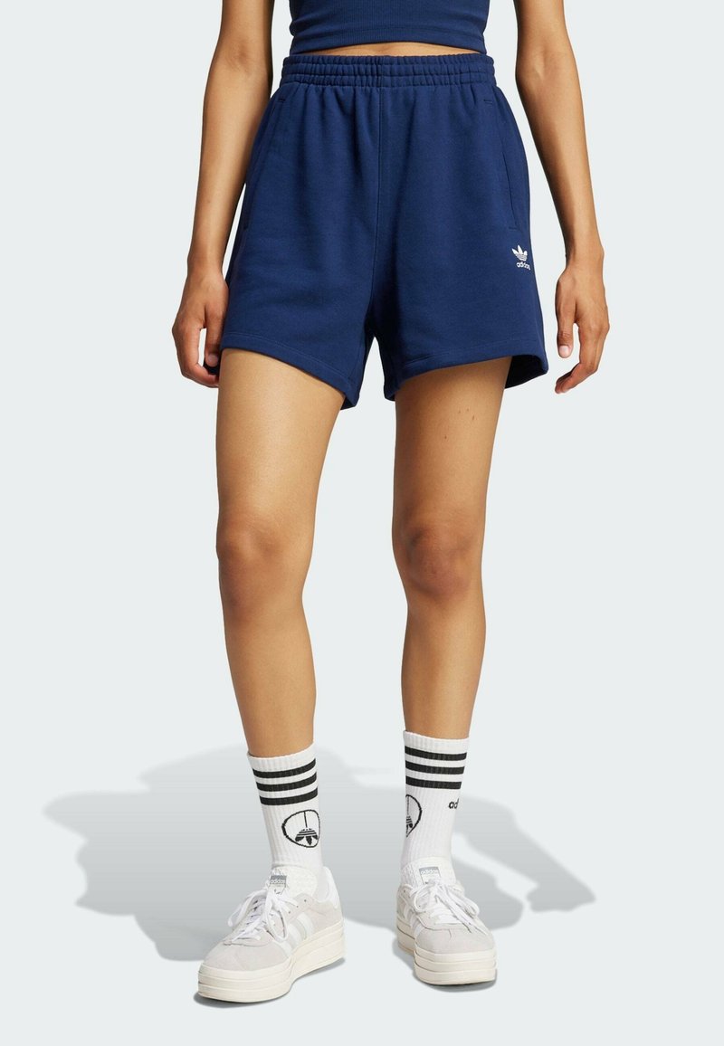 Kvinna som bär marinblå Adidas-shorts med elastisk midja, vita Adidas-strumpor med svarta ränder och vita sneakers som står på en ljus bakgrund.