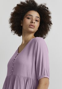 Lavendelfarbenes, strukturiertes Kleid mit fließendem Design, kurzen Puffärmeln und V-Ausschnitt, mit dekorativen Knöpfen an der Vorderseite.