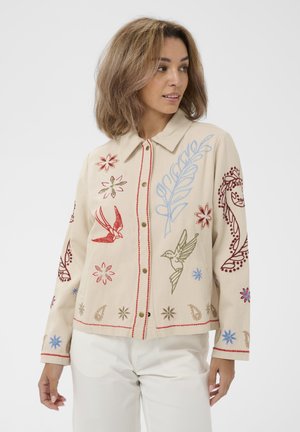 Kobieta w beżowej kurtce z kolorowymi haftowanymi ptakami, kwiatami i liśćmi, w białych spodniach, stojąca na jednolitym tle.