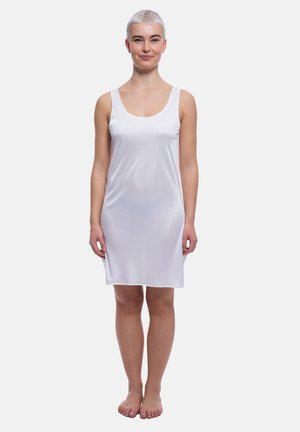 Donna con capelli biondi corti che sta a piedi nudi, indossa un abito bianco senza maniche lungo fino al ginocchio, su uno sfondo semplice.