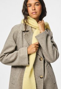 Kvinne iført en beige ullfrakk med store knapper og lommer, som holder et mykt gult skjerf rundt halsen, mot en hvit bakgrunn.