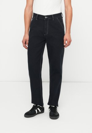 Jeans in denim neri con cuciture bianche a contrasto, design a gamba dritta, chiusura con bottone e passanti per cintura, abbinati a sneakers nere.
