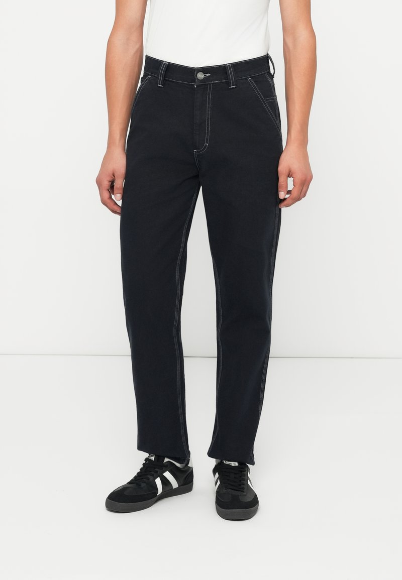 Jeans in denim neri con cuciture bianche a contrasto, design a gamba dritta, chiusura con bottone e passanti per cintura, abbinati a sneakers nere.