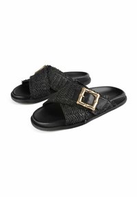 I sandali slip-on neri in tessuto hanno una tomaia testurizzata e un grande dettaglio in metallo dorato. La suola è piatta e realizzata in gomma nera.