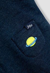 T-shirt in cotone blu navy con una piccola tasca ricamata con un pianeta giallo e un anello blu. L'etichetta riporta "JoJo". Tessuto morbido.