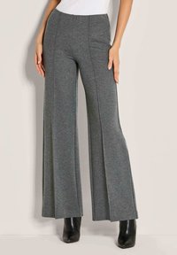 Graue weite Hose mit Vordernähten, kombiniert mit schwarzen Stiefeletten und einem weißen ärmellosen Oberteil, stehend vor einem schlichten hellen Hintergrund.