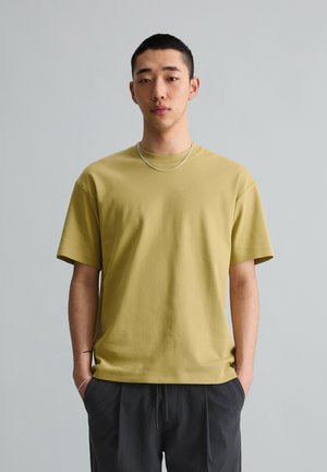 Young man with short black hair wears a yellow short-sleeve t-shirt, silver necklace, and black pants, standing against a plain gray background.