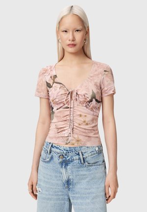 Ung kvinde med langt blondt hår iført en lyserød rynket blomsterprintet bluse med korte ærmer og lyseblå højtaljede jeans.