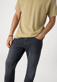 Camisa de algodón verde oliva con cuello redondo y mangas cortas, combinada con jeans de mezclilla gris oscuro de corte relajado y con bolsillos laterales.