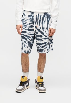 Mand iført sort-hvide tie-dye shorts, hvid langærmet skjorte, hvide sokker og højtop-sneakers i grå, hvid og gul.