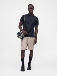 Homme portant un polo bleu marine, un short beige, des chaussettes et chaussures noires, portant un sac à bandoulière à motif avec une sangle sombre sur fond blanc.