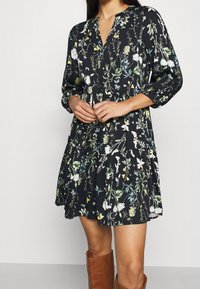 Mujer vestida con un vestido negro con estampado floral y mangas de tres cuartos, con botas marrones hasta la rodilla, con las manos juntas al frente.