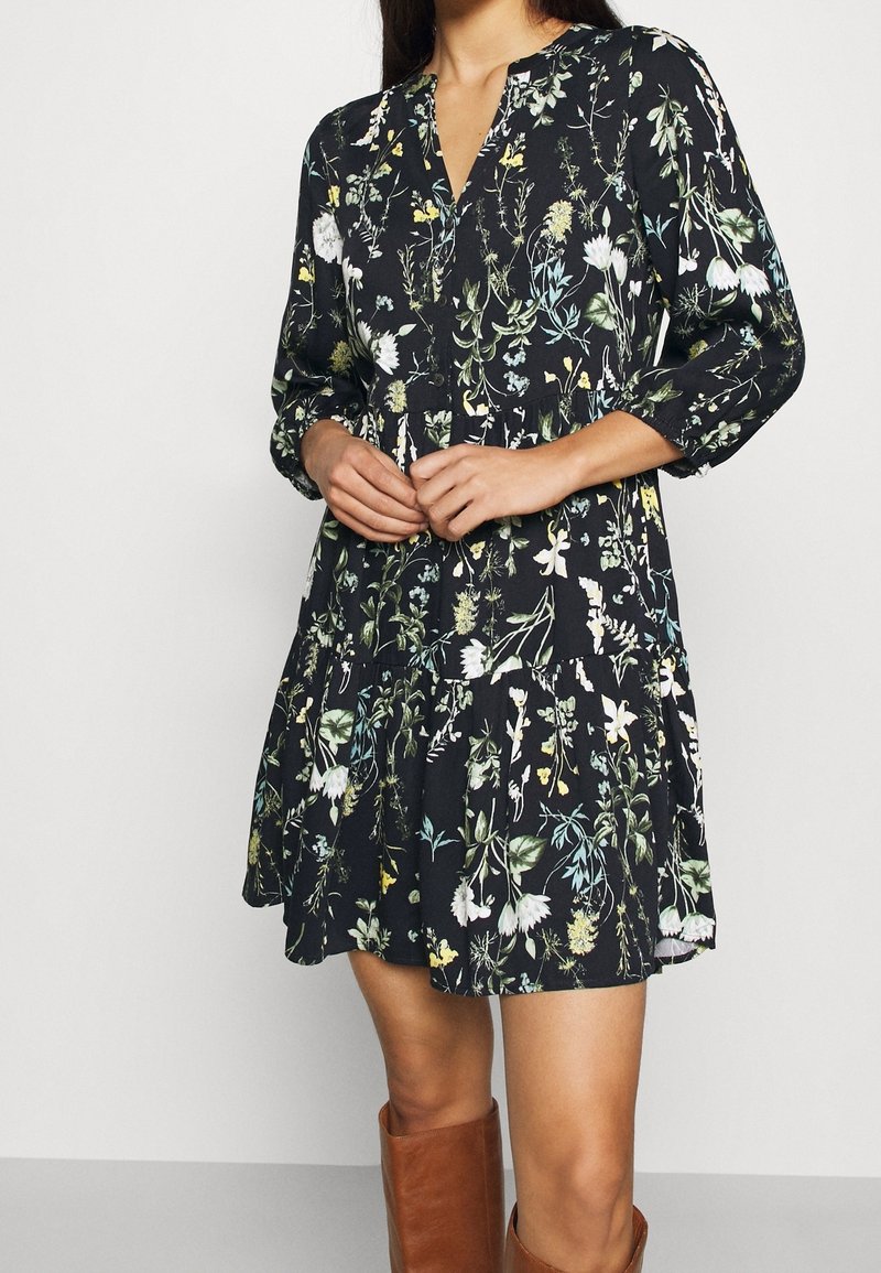 Mujer vestida con un vestido negro con estampado floral y mangas de tres cuartos, con botas marrones hasta la rodilla, con las manos juntas al frente.