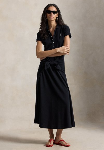 Žena s kučeravými vlasmi v čiernych polo šatách so zapnutými gombíkmi a viazaným opaskom, s čiernymi slnečnými okuliarmi, perlovou náušnicou a červenými sandálmi stojaca s prekríženými rukami.