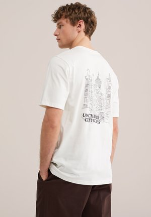 Mladý muž sa pozerá do strán, na sebe má biele tričko s čiernym lineárnym umením vysokých mestských budov a textom „UNCHARTED CITYSCAPES“ na chrbte.