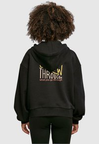 Sudadera negra de gran tamaño con capucha ajustable. Presenta un texto gráfico "CRECE A TRAVÉS de lo que pasas" en colores contrastantes en la parte posterior.