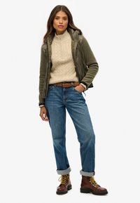 Olijfgroene gewatteerde jas over een crèmekleurige kabelgebreide trui, blauwe opgerolde jeans en bruine veterschoenen met gele veters.