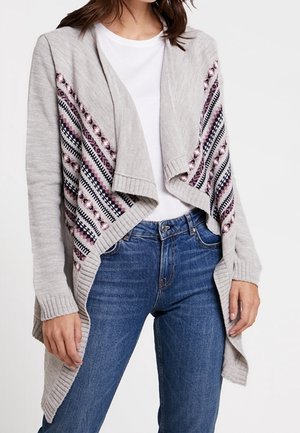 Femme portant un cardigan gris ouvert sur le devant avec des panneaux à motifs roses et noirs, chemise blanche et jean bleu, debout, les bras détendus.