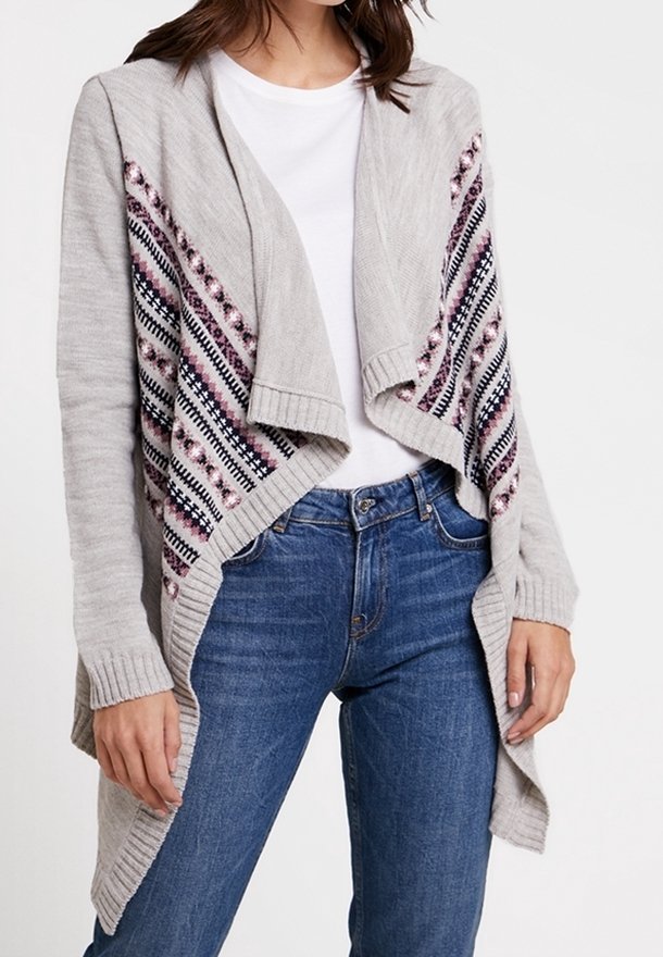 Femme portant un cardigan gris ouvert sur le devant avec des panneaux à motifs roses et noirs, chemise blanche et jean bleu, debout, les bras détendus.