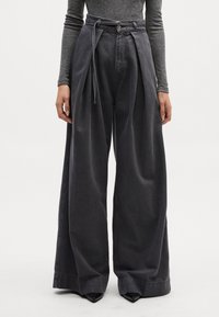 Pantalones de mezclilla oscuros de pierna ancha con plisados, cinturilla con hebilla y una textura suave. Presenta un cierre de botón y un diseño de cintura alta.