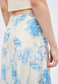 Λευκή φούστα με μπλε λουλουδάτο σχέδιο, φορεμένη από άτομο με ακάλυπτη κοιλιά και ανοιχτόχρωμο δέρμα, που στέκεται μπροστά σε απλό φόντο.