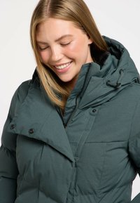 Chaqueta de plumas aislante verde con capucha, que presenta un cierre de cremallera en la parte delantera, acentos de botones y una tela texturizada. Se muestra con un cuello en ángulo y detalles de bolsillo.