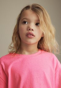 Světle růžové bavlněné tričko s kulatým výstřihem, charakterizované jemnou texturou a volným střihem. Vlnité blond vlasy částečně rámují obličej.