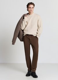 Beiger Strickpullover, braune Hose, schwarze Schuhe und eine graue Jacke, die über der Schulter hängt. Schlichte Texturen, neutrale Farben und maßgeschneiderte Passform.