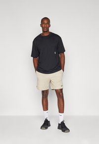 Black cotton t-shirt with short sleeves, paired with beige shorts. Features a small pocket and black athletic shoes with white socks.